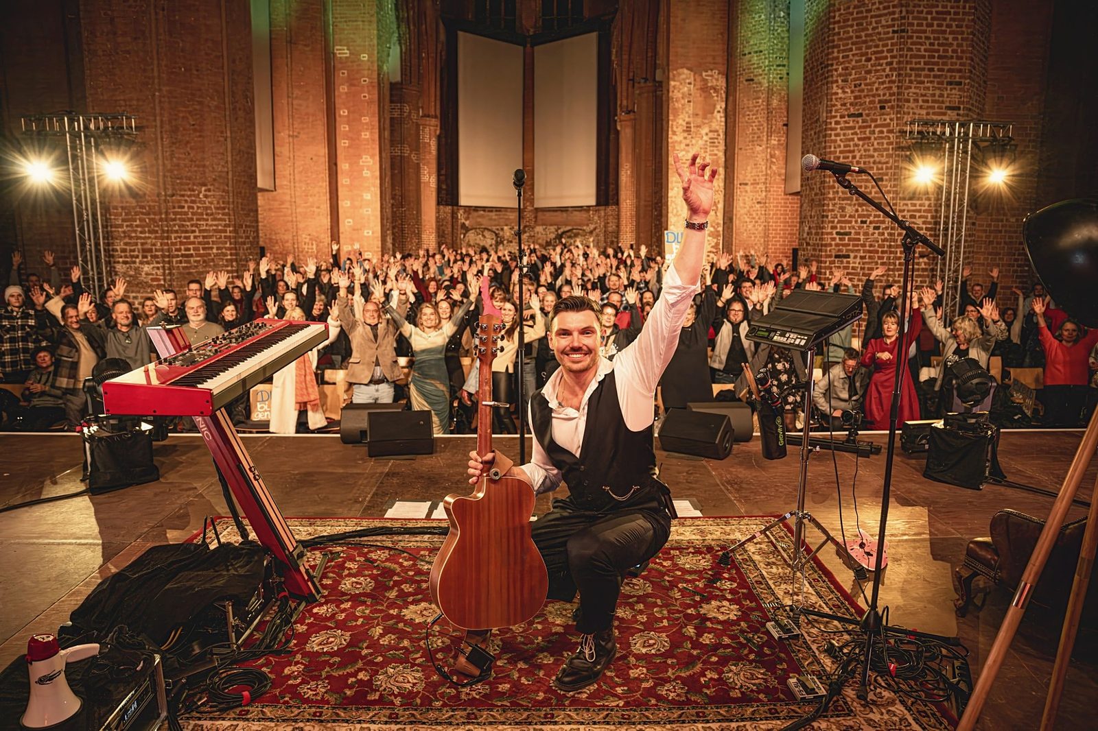 ÖXL kniet lachend mit Gitarre vor der vollen St. Georgenkirche Wismar, das Publikum hebt die Hände